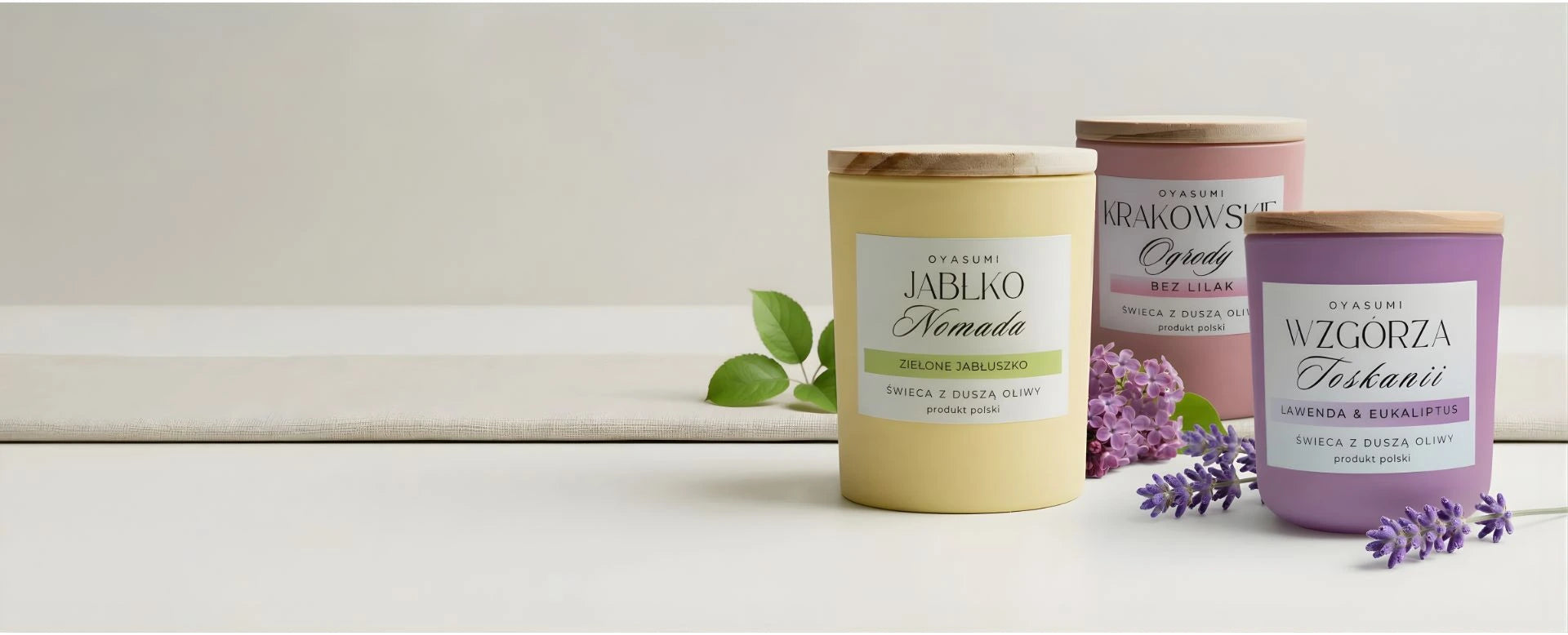 Kolekcja wiosennych świec z wosku oliwnego Oyasumi: Jabłko Nomada, Krakowskie Ogrody oraz Wzgórza Toskanii. Polskie świece zapachowe o aksamitnej strukturze w pastelowych opakowaniach z drewnianymi wieczkami, otoczone kwiatami bzu i lawendy.