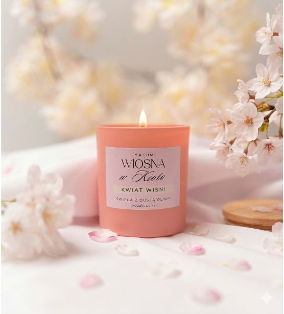 Różowa świeca z wosku oliwnego Wiosna w Kioto od Oyasumi, zapalona, umieszczona na białym materiale w otoczeniu kwitnącej wiśni. Elegancki baner kolekcji wiosennej w jasnej, pastelowej kolorystyce.