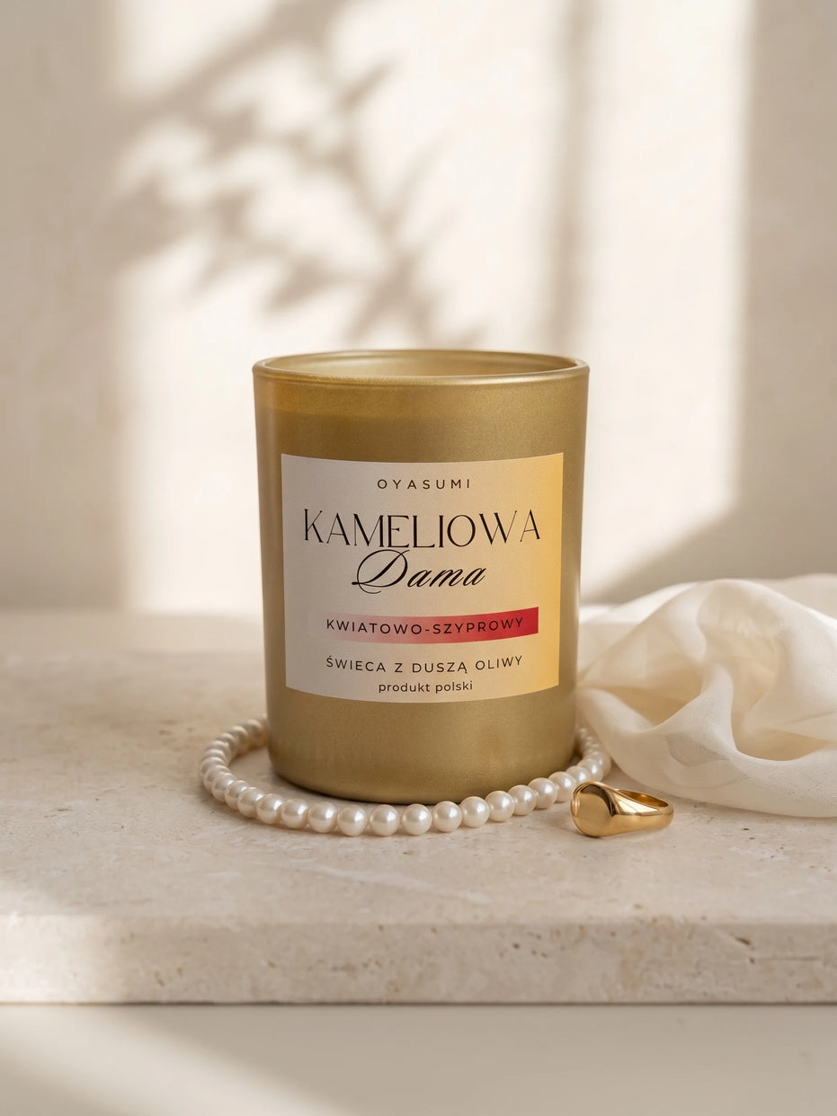 Świeca Kameliowa Dama wyeksponowana na jasnym kamieniu obok sznura pereł, złotego sygnetu i białej tkaniny z delikatną grą cieni.
