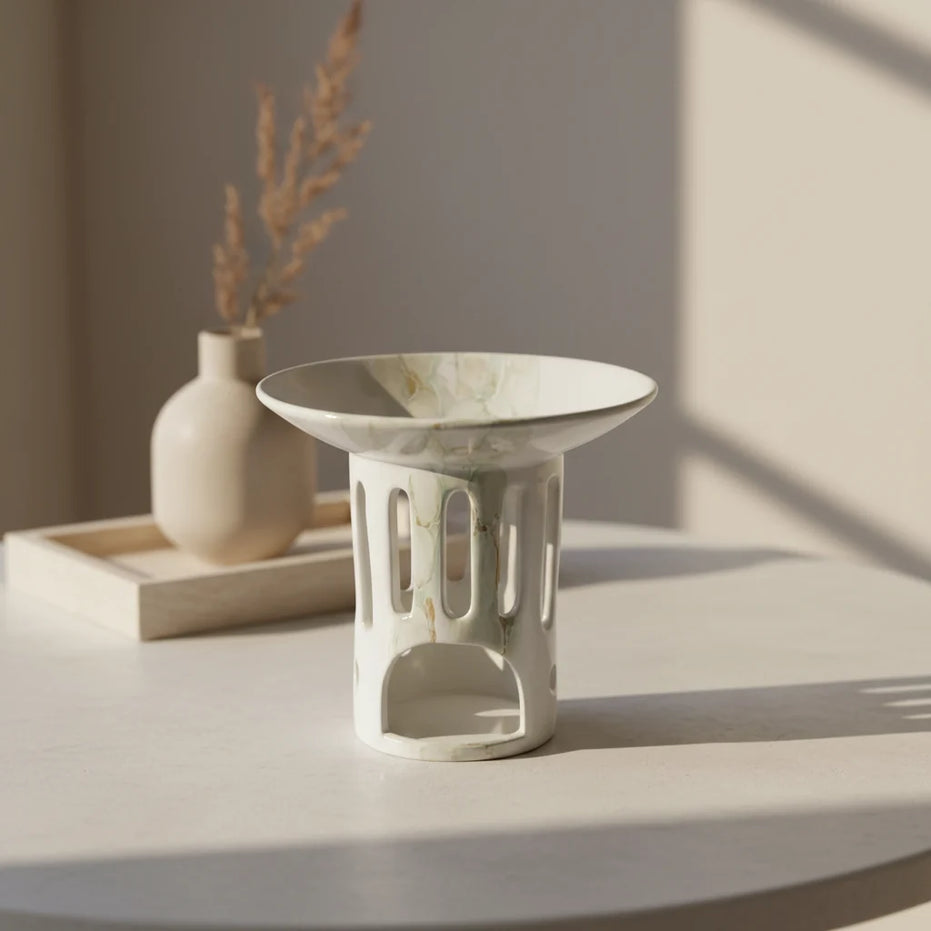 Ceramiczny kominek do wosków zapachowych OYASUMI w białym marmurkowym wykończeniu na okrągłym stoliku z dekoracyjnym wazonem.