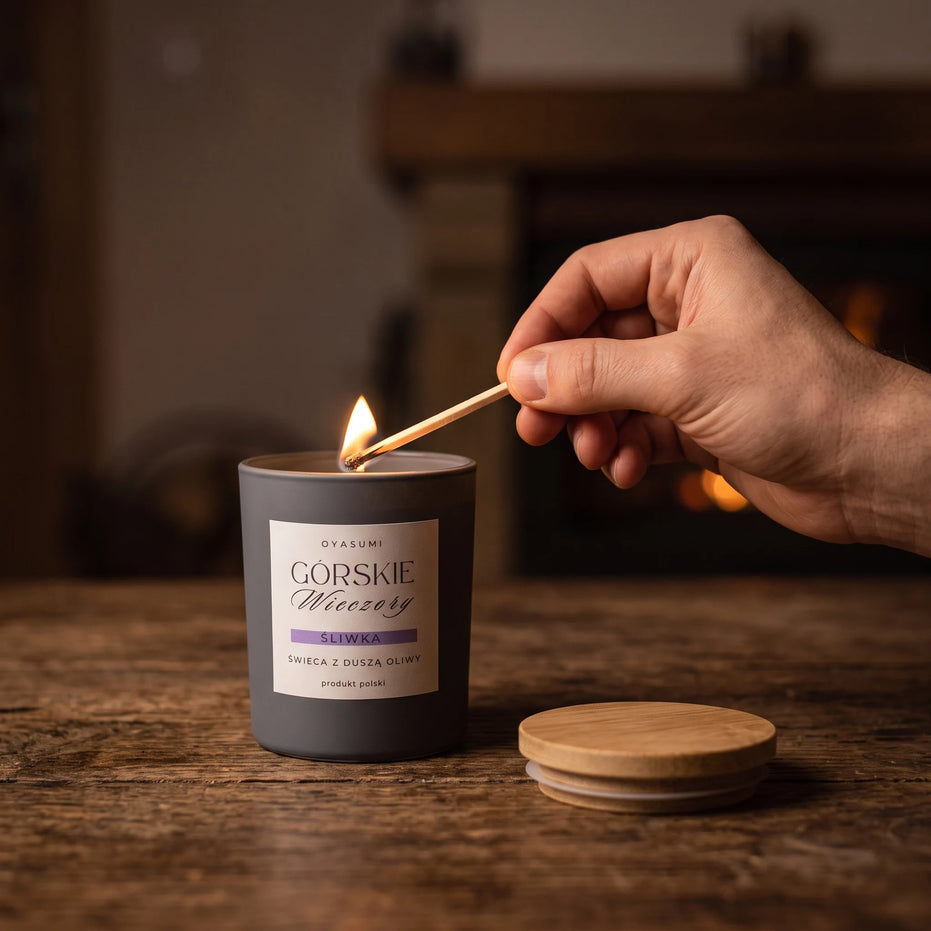 Dłoń zapalająca świecę zapachową Górskie Wieczory oyasumi zapałką przy kominku – śliwkowy zapach do domu, wosk oliwny, produkt polski