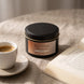 Świeca zapachowa OYASUMI Rzymskie Espresso 90 ml z książką i filiżanką kawy w ciepłej, domowej aranżacji.