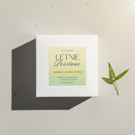 Białe pudełko wosku zapachowego Letnie Positano marki Oyasumi obok zielonego liścia bambusa na jasnym tle.