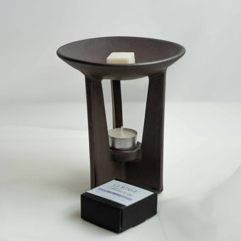 £Wysoki, ceramiczny kominek zapachowy Star firmy Oyasumi. Na górze szerokiej misy kostka wosku oliwnego o zapachu śliwki. Przed kominkiem czarne pudełko z woskami oliwnymi Górskie Wieczory.